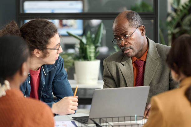 Portrait of Black senior businessman looking over glasses at young colleague while collaborating during business meeting in inclusive office